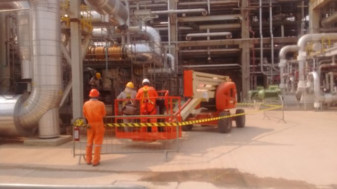 Trabajadores en un sitio industrial rodeados de tuberías y maquinaria pesada.